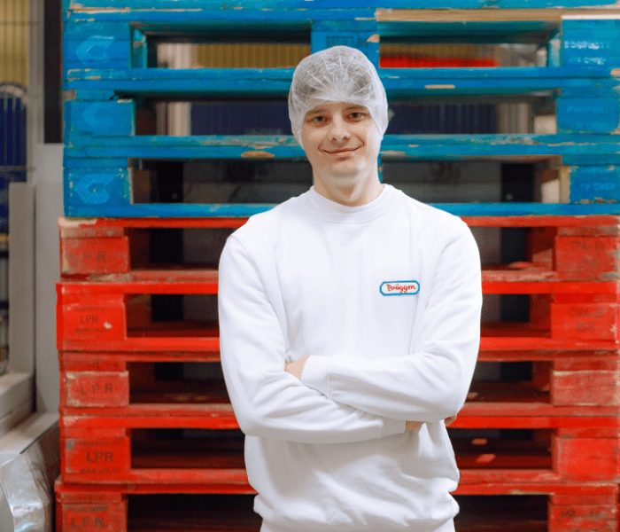 Ein lächelnder Mitarbeiter der H. & J. Brüggen KG in weißer Arbeitskleidung und Haarnetz vor einem Lager mit Holzpaletten.