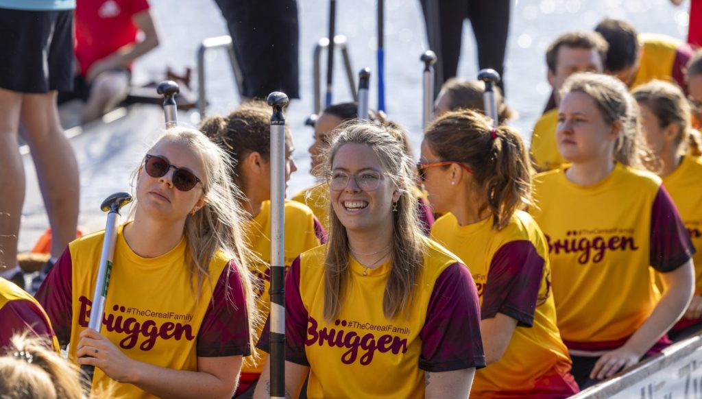 Eine Gruppe von Frauen in gelb-weinroten Brüggen-T-Shirts hält Paddel in einem Boot, während eine von ihnen fröhlich lächelt.