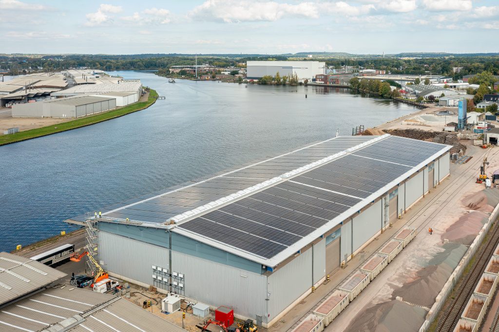 Das Bild zeigt eine Luftaufnahme der Logistikhalle der Firma H. & J. Brüggen KG, auf dem Dach sieht man die Photovoltaikanlage, im Hintergrund sieht man ein weiteres Werk der Firma Brüggen am Wasser