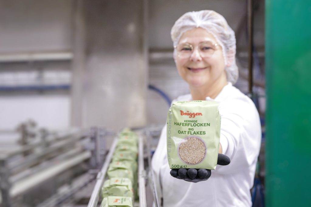 Das Bild zeigt eine lächelnde Frau im Werk einer Haferflockenproduktion mit einer Haferflockenpackung in der Hand