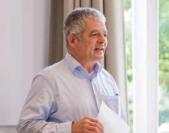 Ein Mann, Hanno Brüggen, hält eine Rede, während er vor jungen Personen steht. Er hält ein weißes Blatt Papier in der Hand.