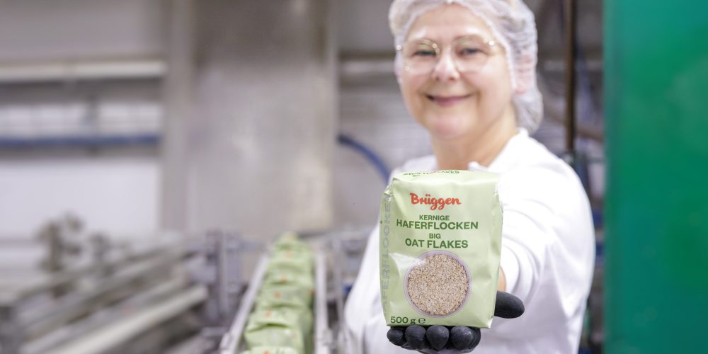 Das Bild zeigt eine lächelnde Frau im Werk einer Haferflockenproduktion mit einer Haferflockenpackung in der Hand