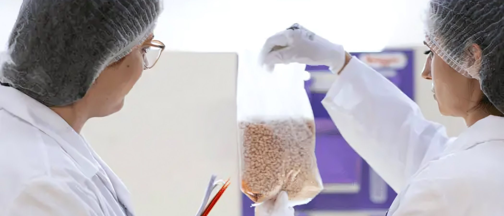 Zwei Mitarbeiterinnen der H. & J. Brüggen KG im weißen Kittel und mit Hygienekappen bei der Qualitätskontrolle von Müsli-Rohstoffen im Labor.